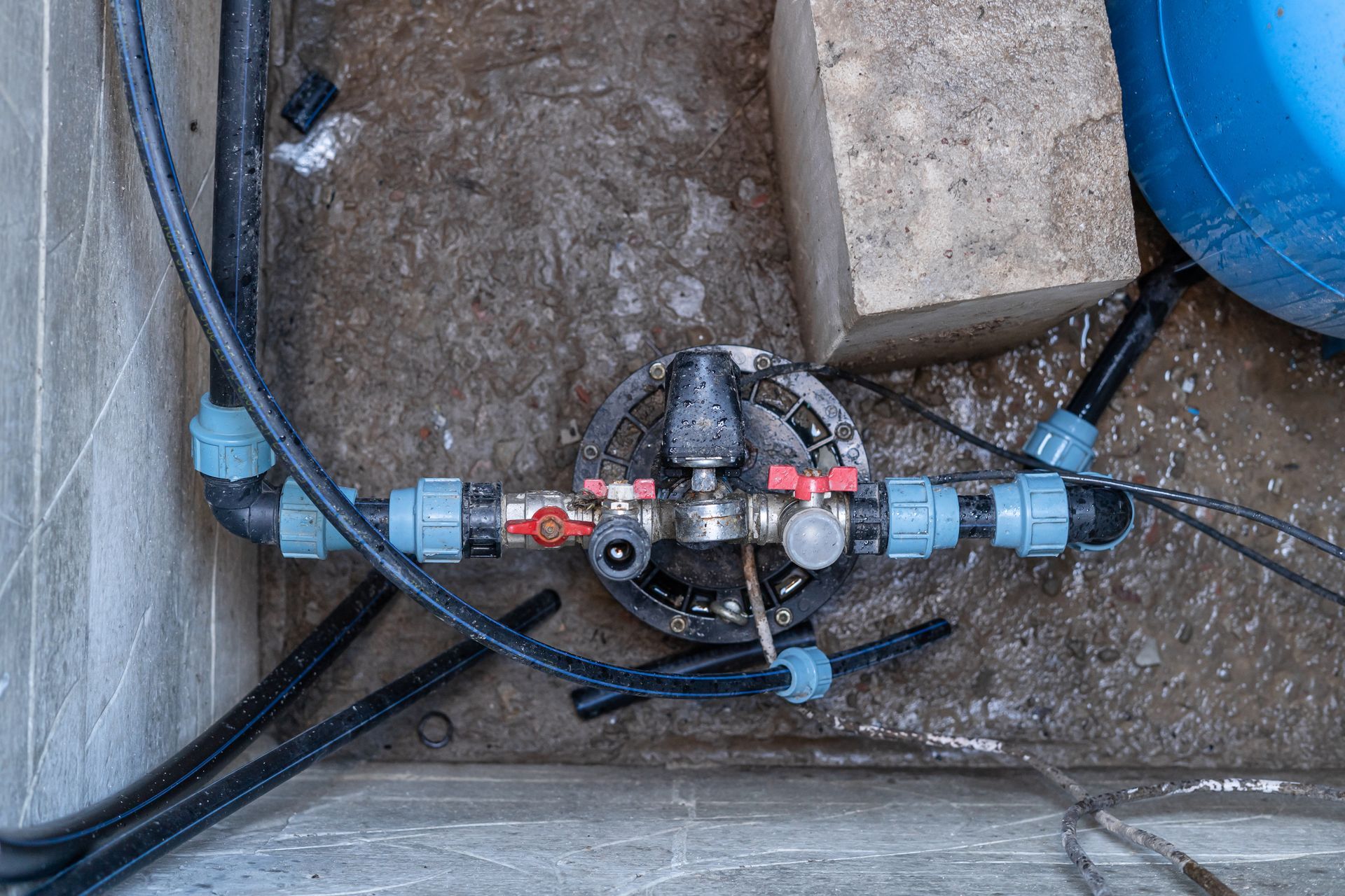 An irrigation system manifold with black pipes and blue connectors mounted on a dirt ground surface next to a concrete block.