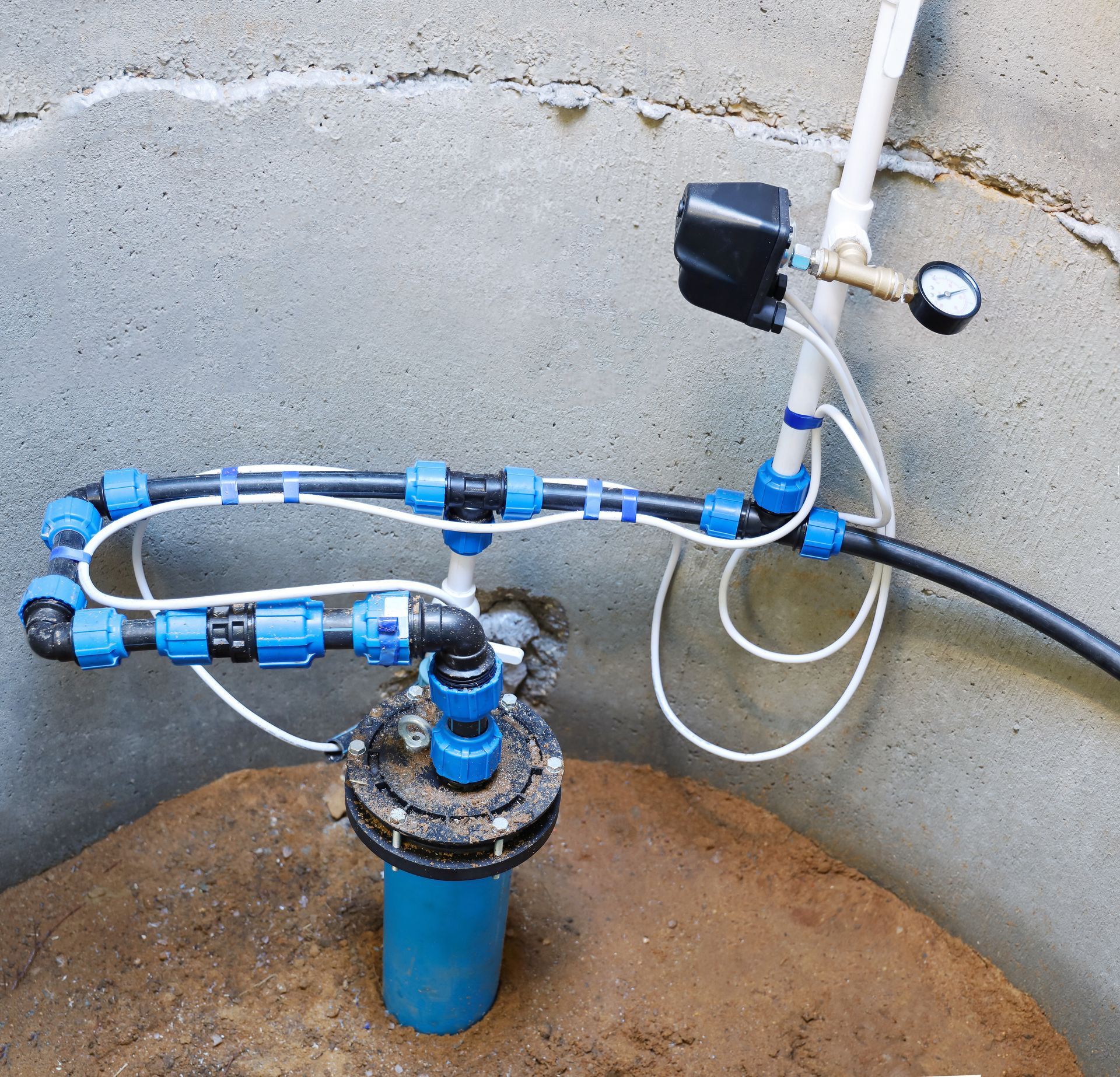 Water well pump assembly with blue plastic pipes, a pressure gauge, and electrical wiring inside a concrete enclosure.