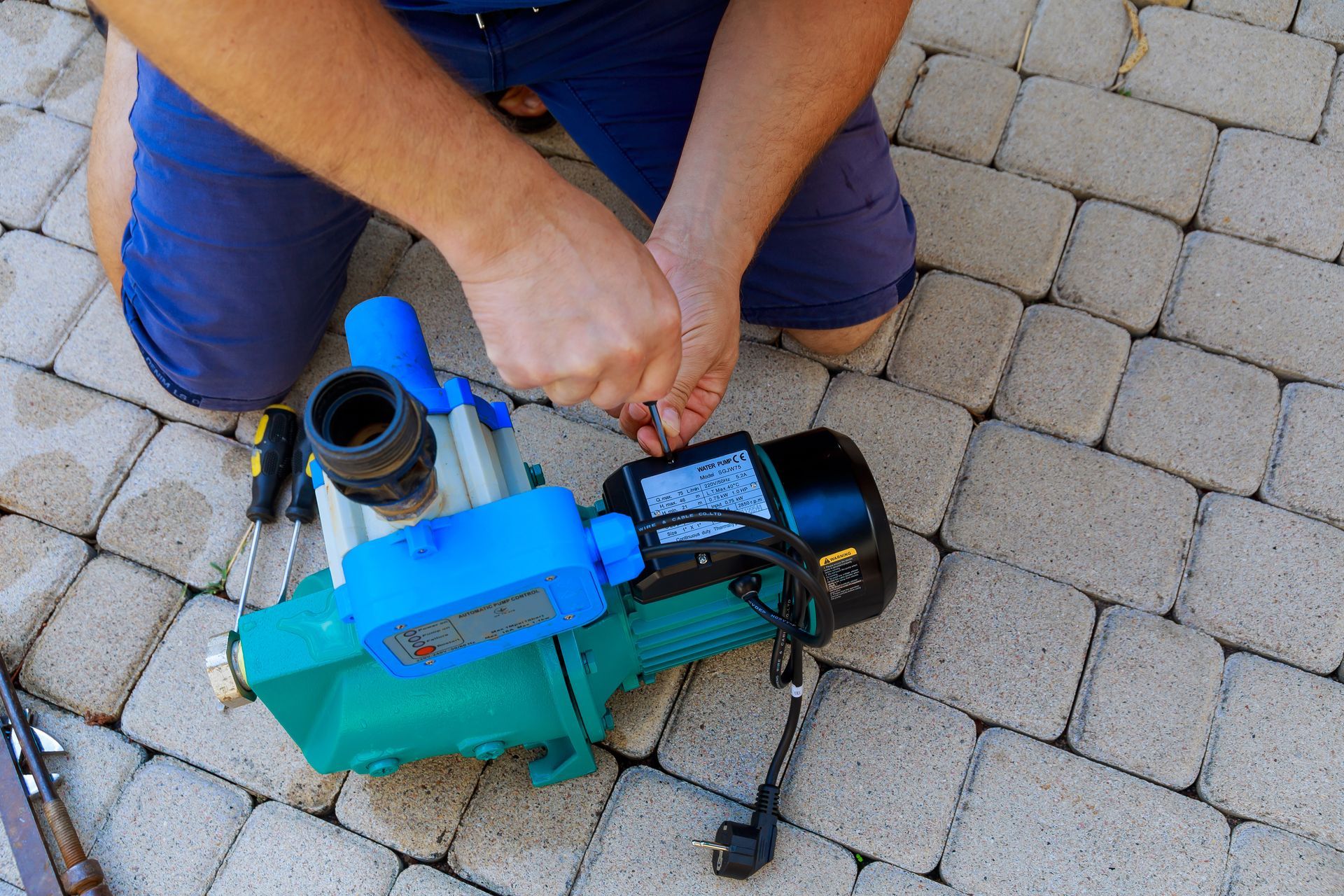 A person works on a teal electric water pump with a screwdriver, kneeling on a paved surface.