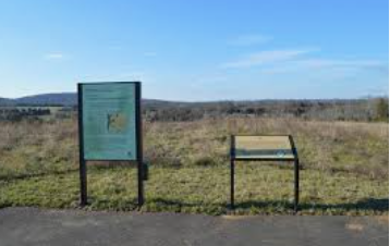 Trail sign and bench beside a paved path in a grassy open field under a blue sky