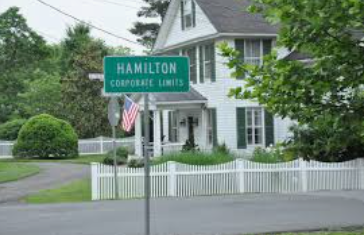 Hamilton corporate limits sign in front of a white house with a picket fence and American flag