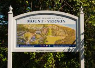 A rectangular sign for George Washington’s Mount Vernon featuring a map of the estate on a white post frame among trees. A rectangular sign for George Washington’s Mount Vernon featuring a map of the estate on a white post frame among trees.
