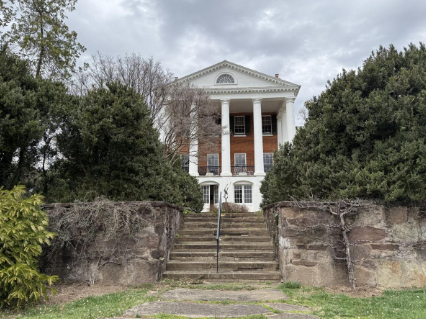 A large, white-pillared historic brick mansion viewed from the bottom of stone steps surrounded by evergreen bushes. A large, white-pillared historic brick mansion viewed from the bottom of stone steps surrounded by evergreen bushes.