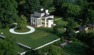Aerial view of a white house with a green lawn, circular driveway, and surrounding trees