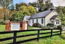 White church with steeple behind a black fence and sign, surrounded by trees and greenery