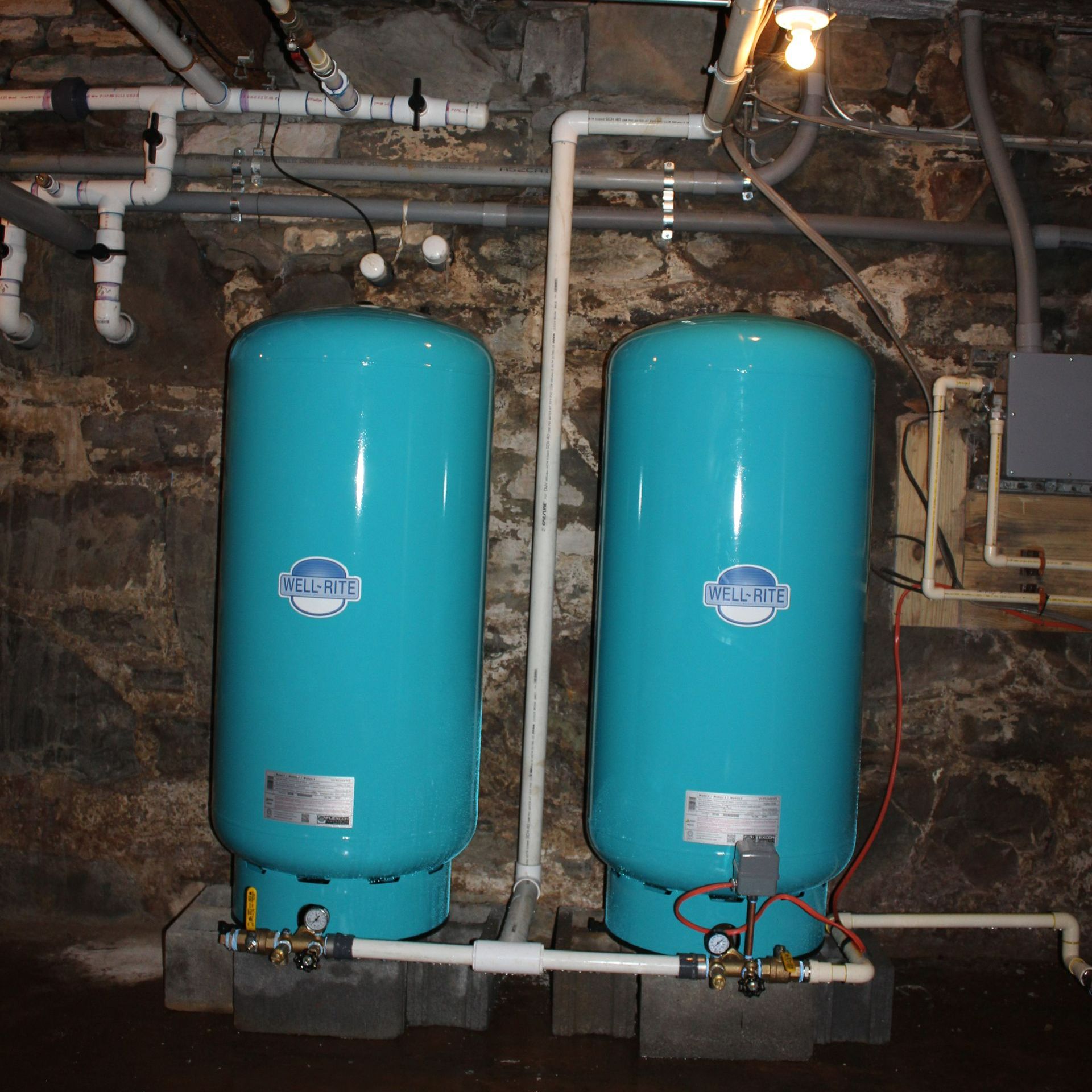Two tall, blue water tanks with white pipes against a stone wall in a basement.
