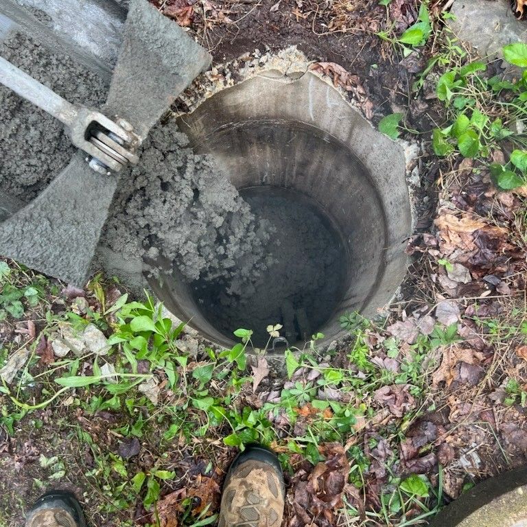 Hole in the ground with gray soil being scooped out by a metal tool. Green plants surround the hole.