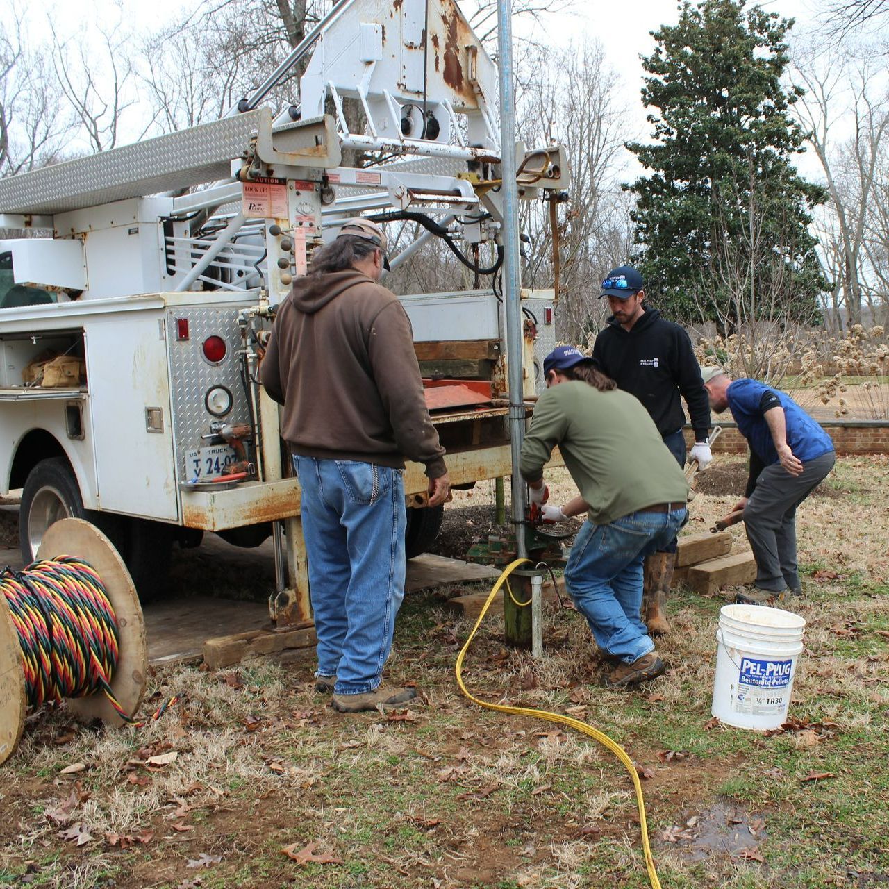 Workers installing a well pump near a utility truck and a cable reel outdoors.