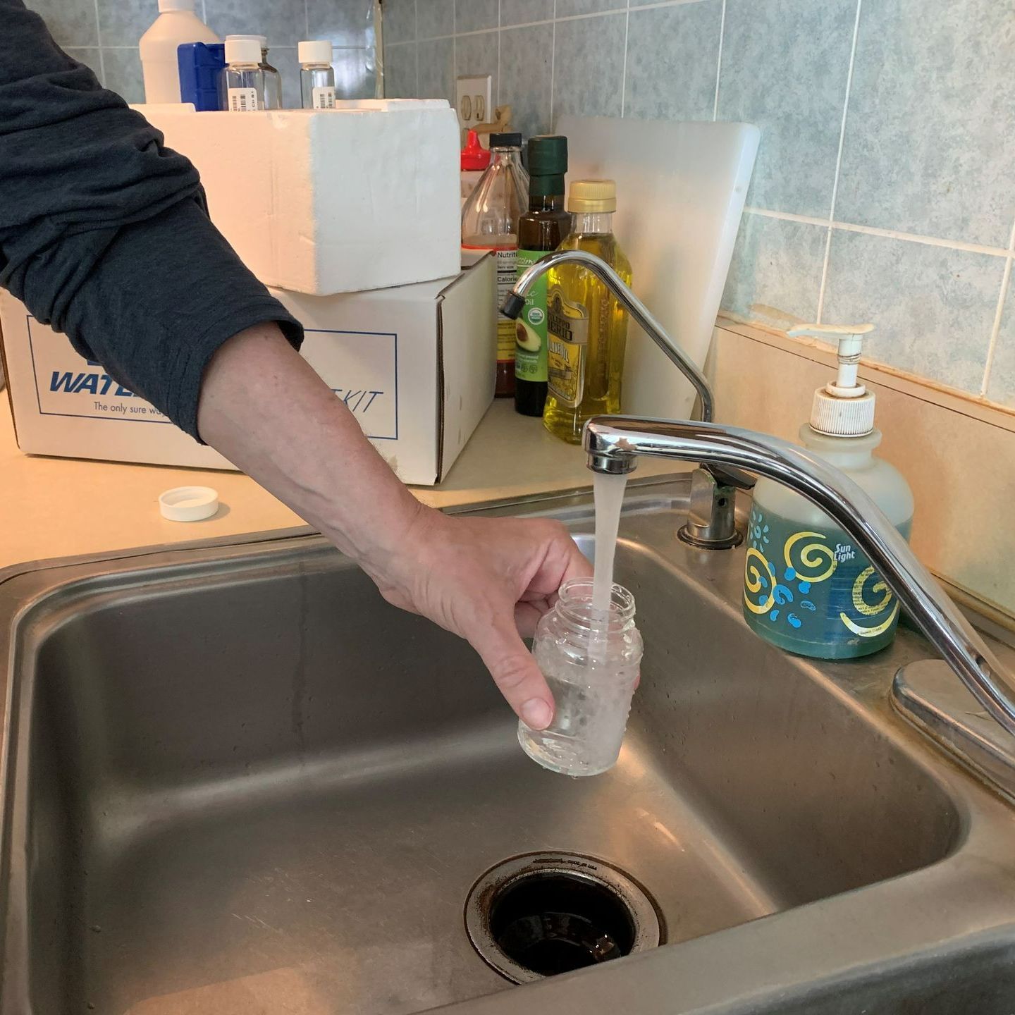 Person filling a small clear bottle with water from a kitchen faucet in a stainless steel sink.