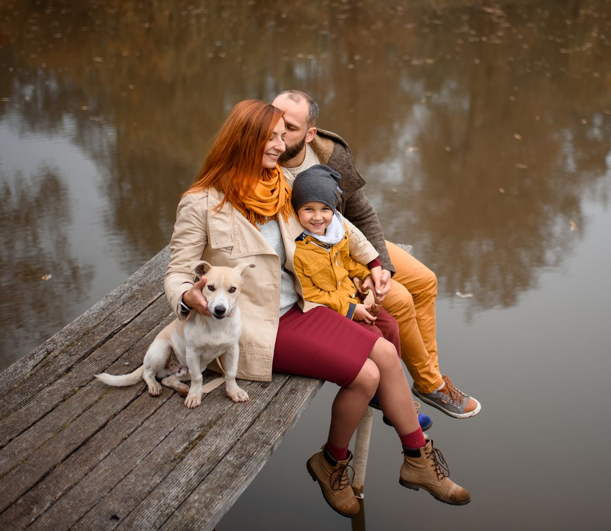 Une famille avec un chien est assise sur un quai, profitant de l'automne au bord de l'eau ; une femme aux cheveux roux, un enfant en veste jaune.