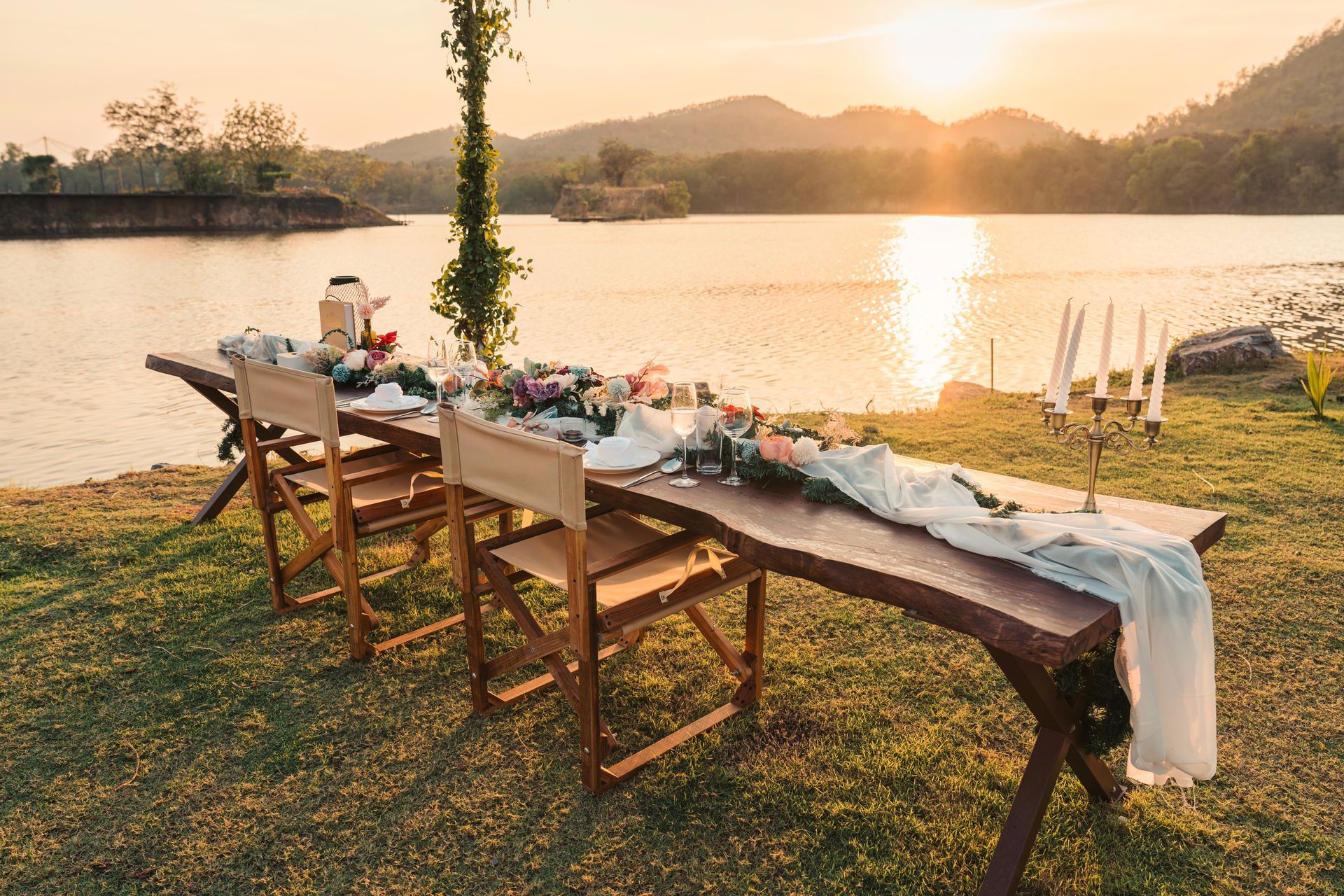 Longue table en bois dressée pour un repas, avec vue sur un lac au coucher du soleil.