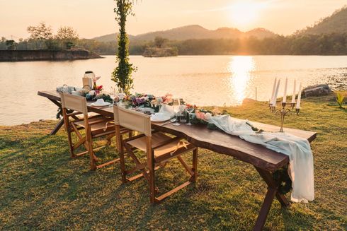Longue table en bois dressée pour un repas, avec vue sur un lac au coucher du soleil.