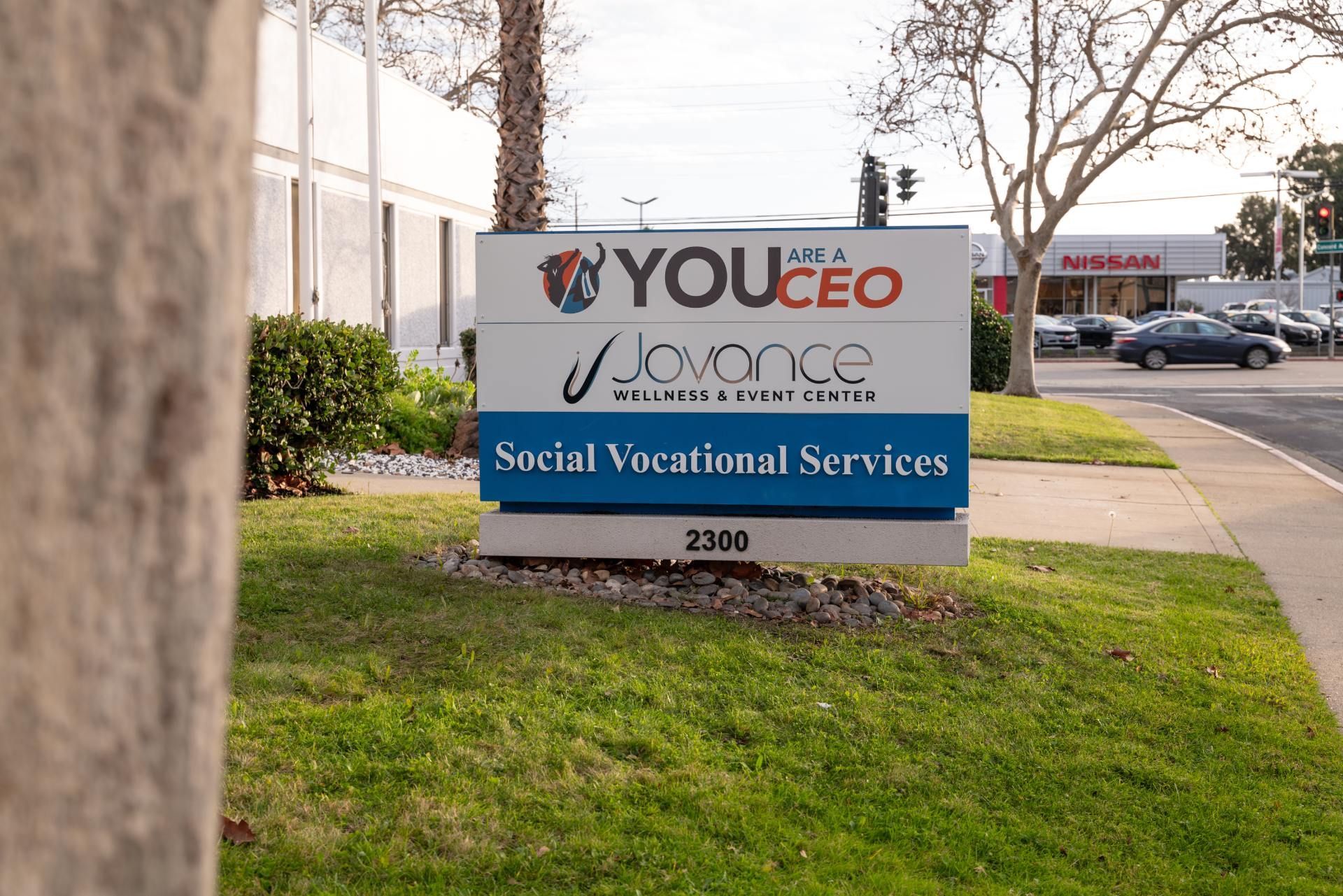 A sign for you ceo is sitting in the grass in front of a building.
