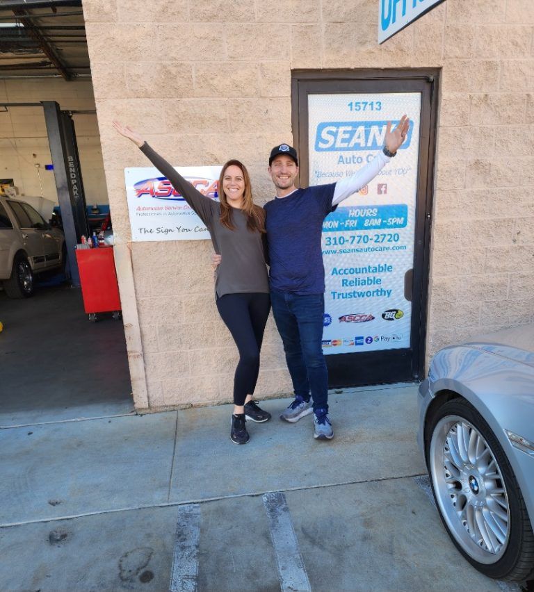 A man and a woman are standing in front of a sean auto shop - Sean's Auto Care
