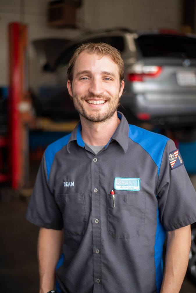 A man in a gray and blue shirt is standing in front of a car in a garage - Sean's Auto Care