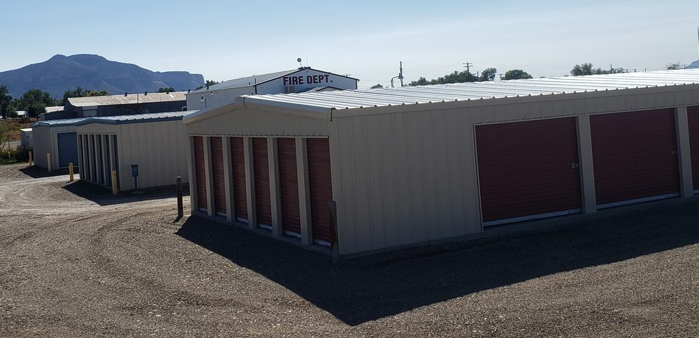 Storage units, beige metal buildings with red doors, gravel lot. Mountains in the background on a sunny day.
