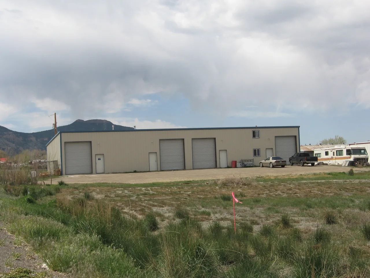 A light beige industrial building with four garage doors, in front of a grassy field with a mountain in the background.