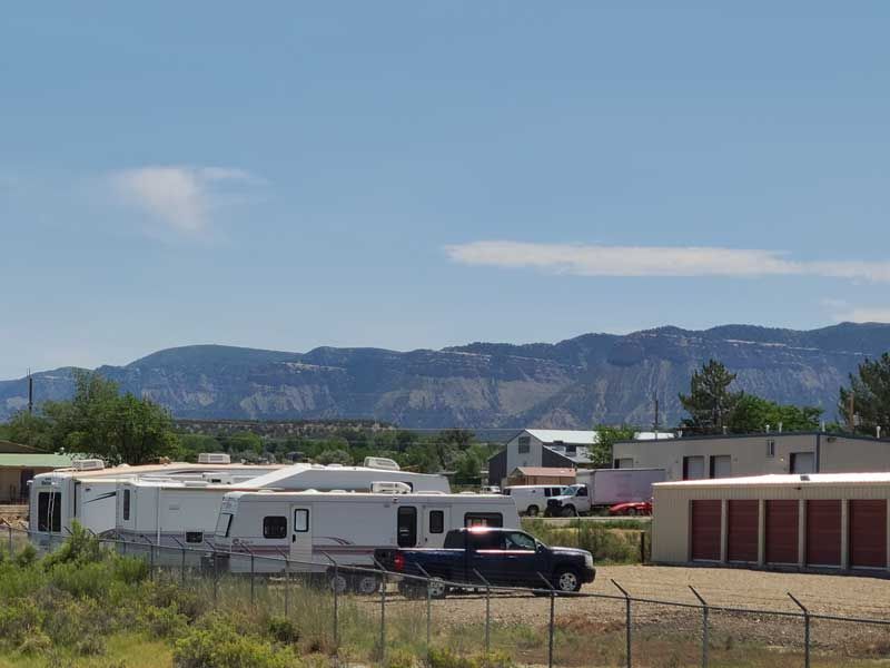 RVs parked near storage units, with a truck, fence, and mountains in the background. Bright sunny day.