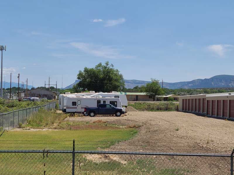 A parked RV is next to a truck. Storage units are visible on the right with mountains in the background under a blue sky.