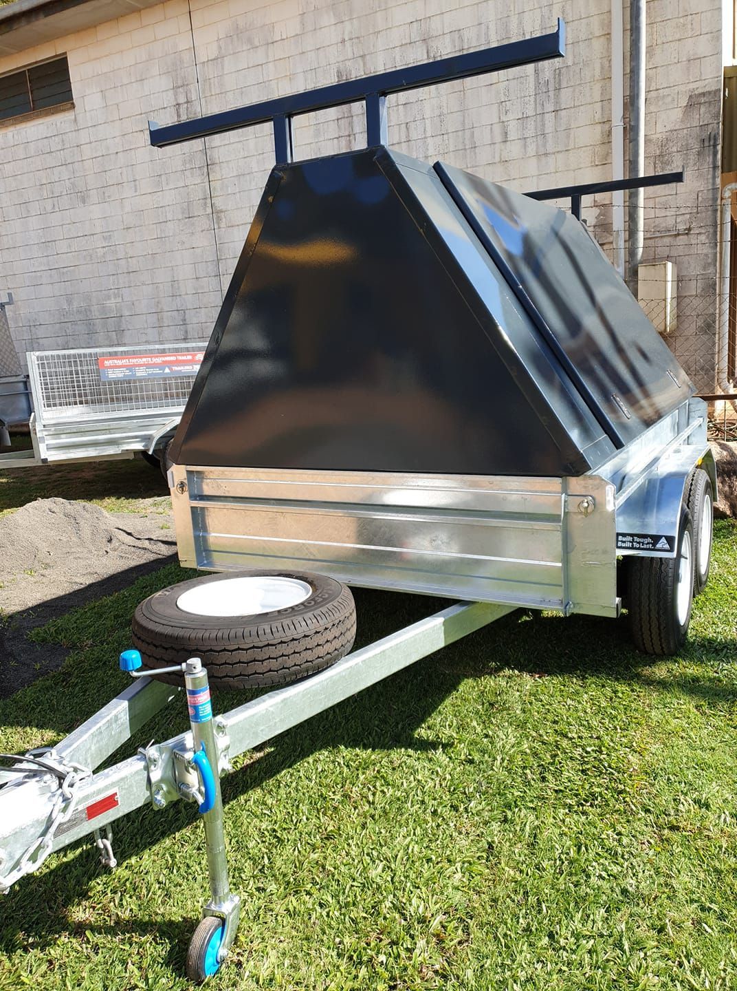 A Trailer is Parked in Front of a Wooden Fence — Tolga Traders in Tolga, QLD Silver utility trailer with black cover and spare tire on green grass. — Tolga Traders in Tolga, QLD