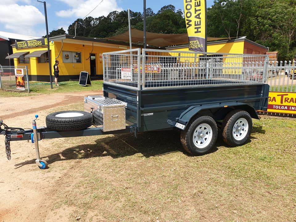 Dark blue trailer with white wheels, spare tire, and metal cage, parked outside a yellow building. — Tolga Traders in Tolga, QLD