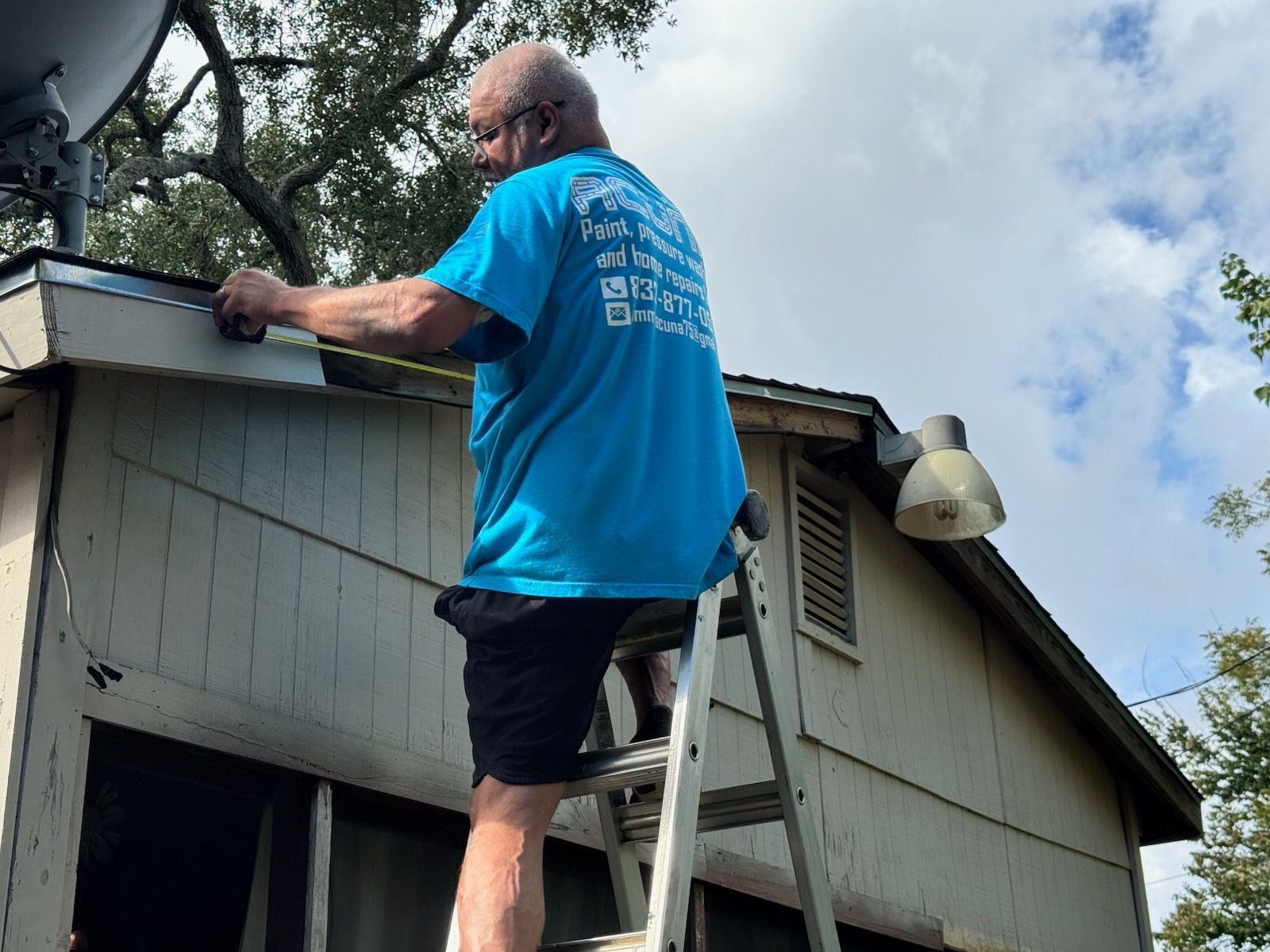 Man on roof cleaning gutters with a ladder beside a gray house under a cloudy sky