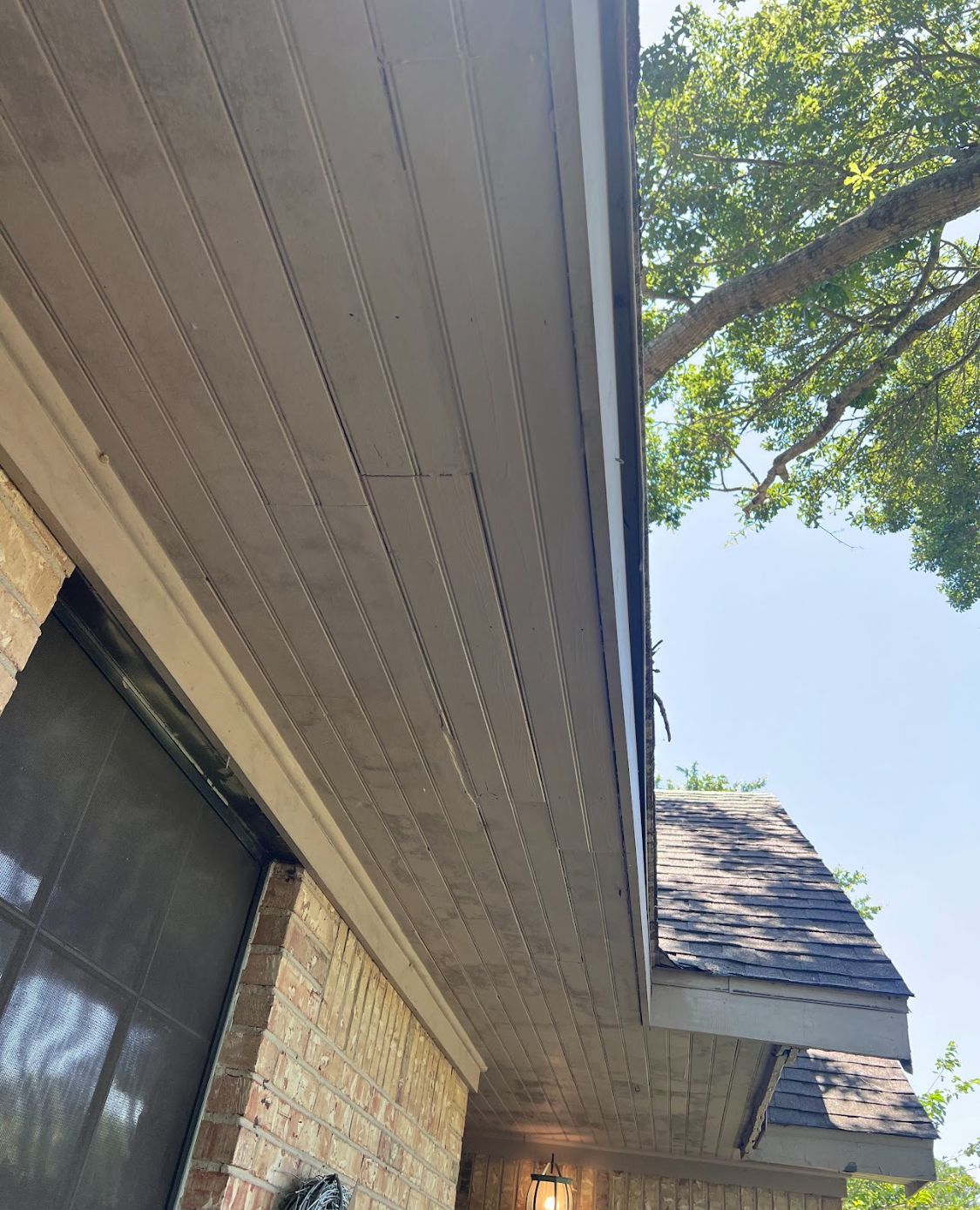 Exterior view of a house roofline with tan brick siding and leafy trees against a blue sky