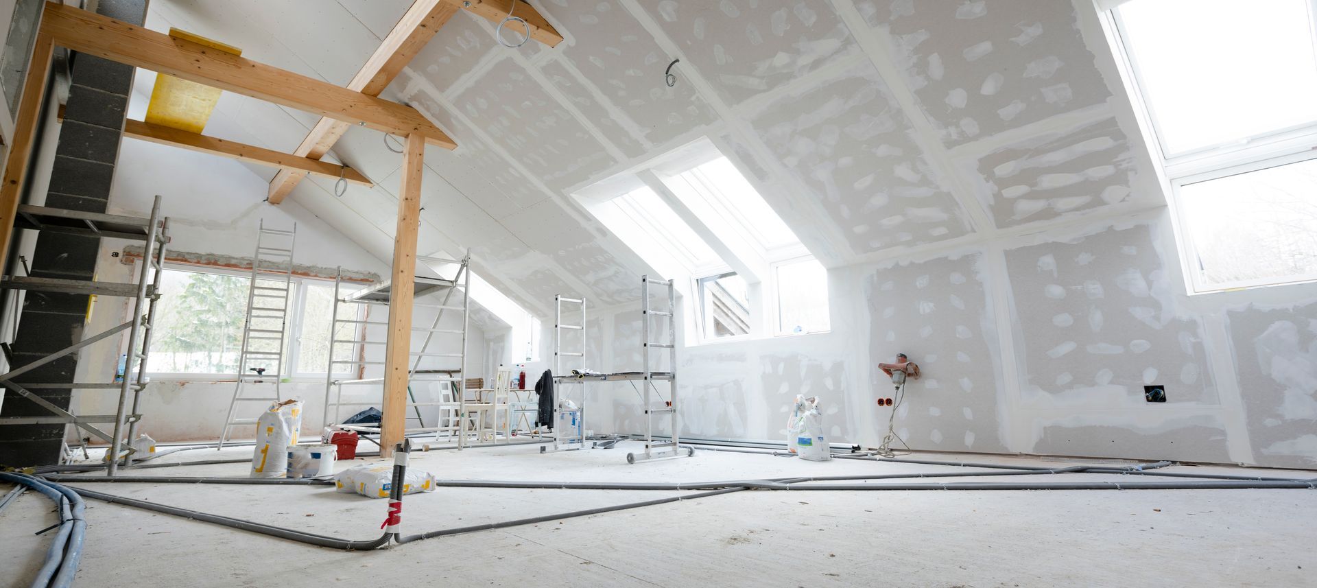 Bright unfinished room with drywall, ladders, scaffolding, and a worker near large windows during renovation