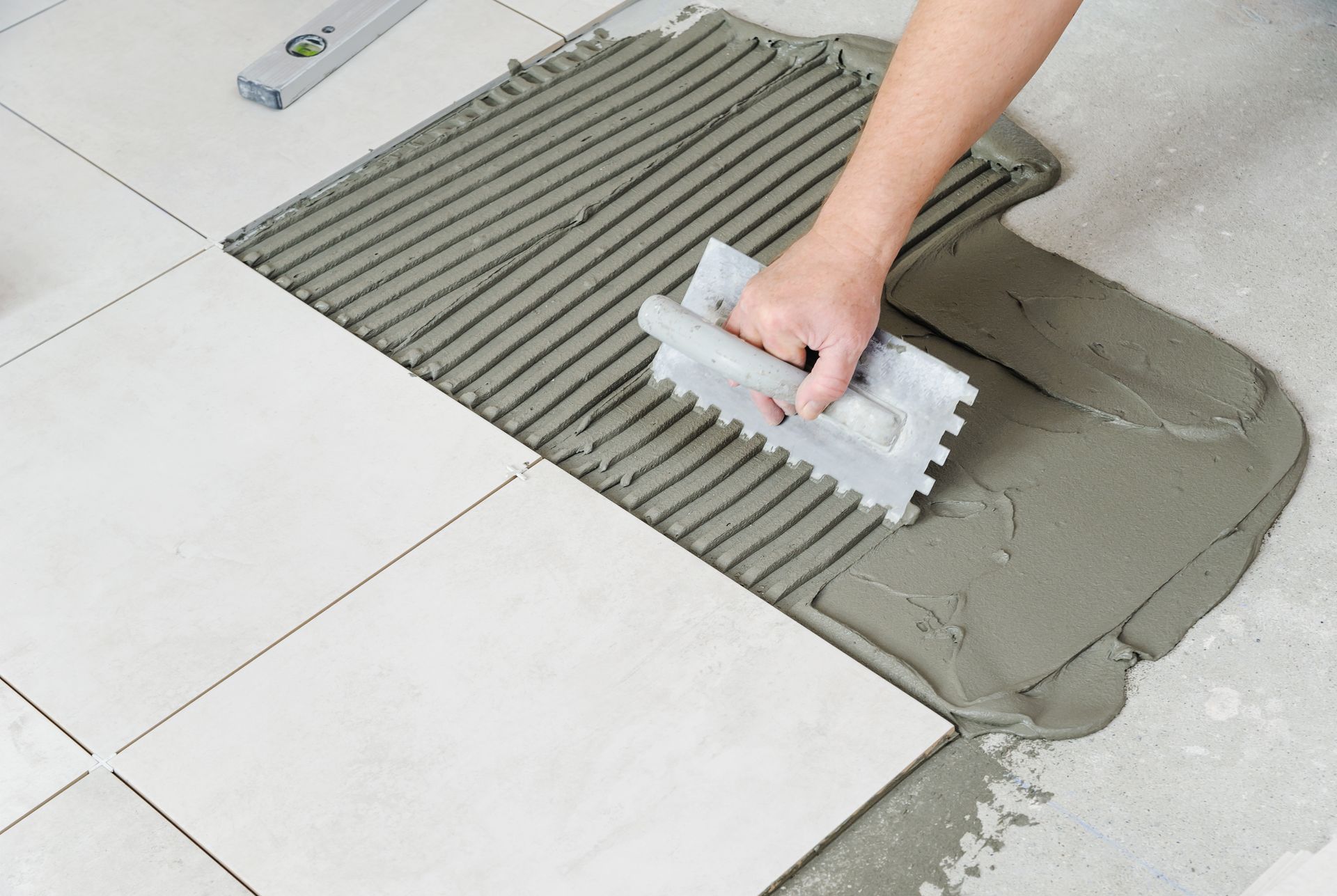 Hand trowel spreading gray tile adhesive on a tiled floor