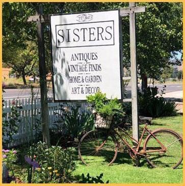 A bicycle is parked in front of a sign for sisters antiques.