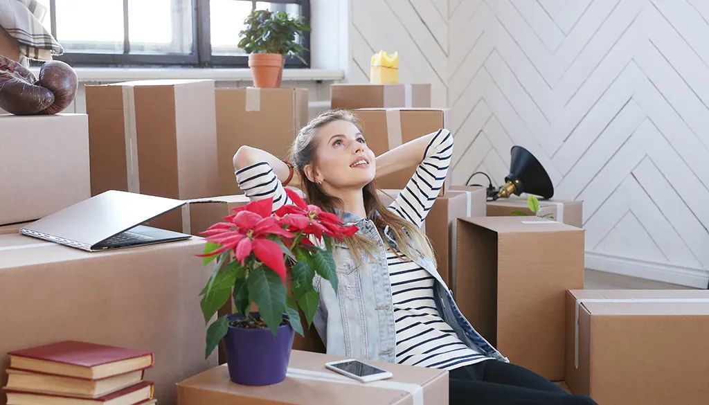 A woman is sitting in a living room surrounded by cardboard boxes.