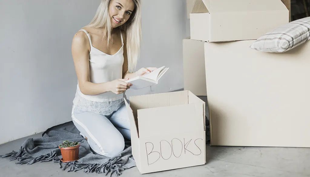 A woman is sitting on the floor next to a cardboard box that says books.