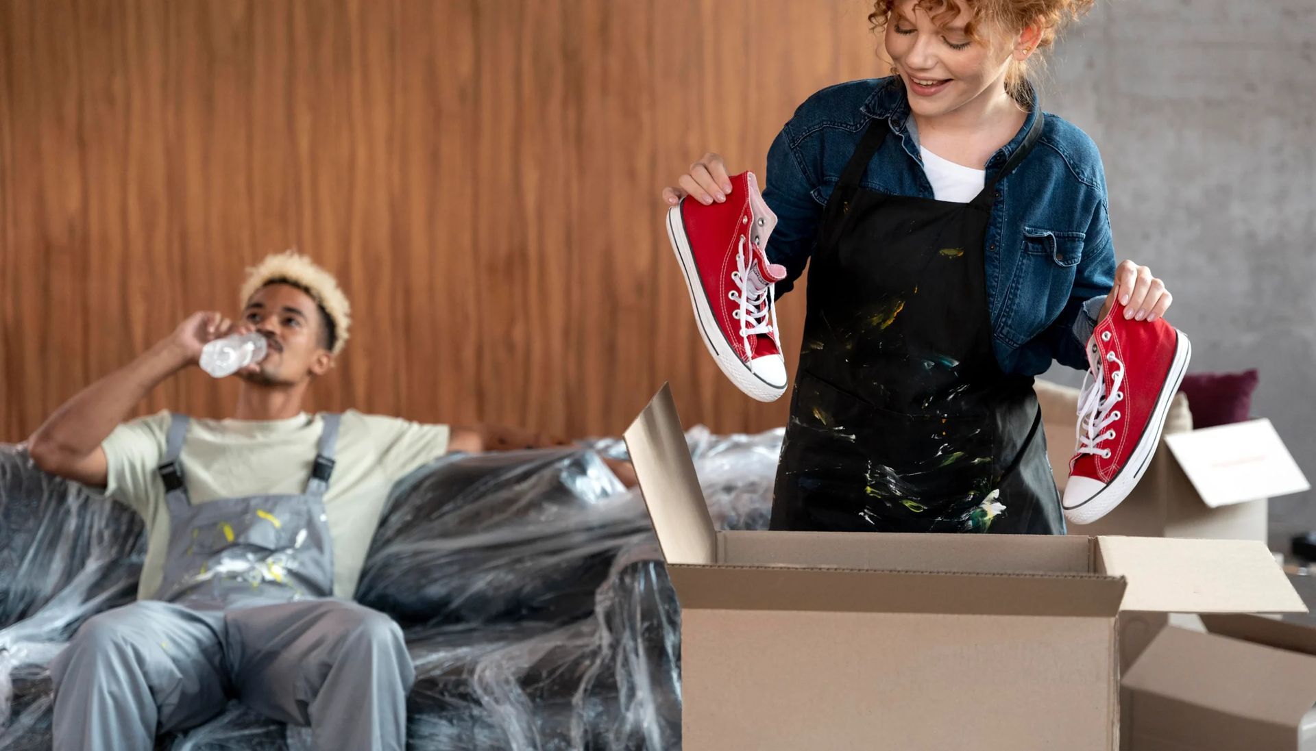 A man is sitting on a couch while a woman is holding a pair of red sneakers.