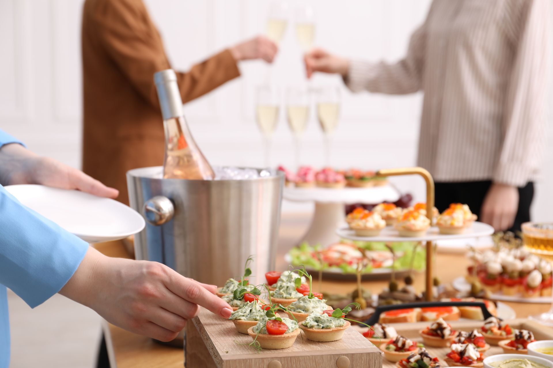 People at a party, reaching for appetizers and toasting with champagne; food display on a table.