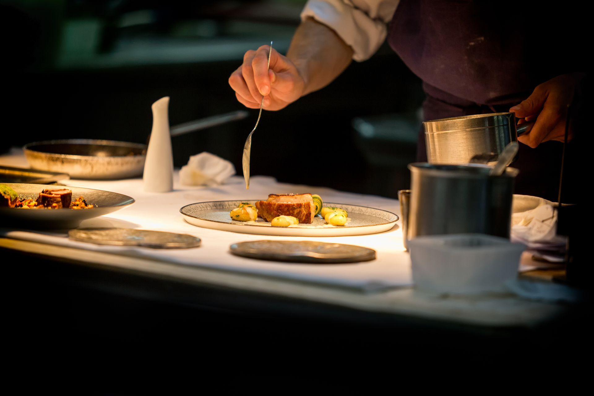 Chef garnishing a plated meal with sauce; dim lighting, stainless steel and white table.