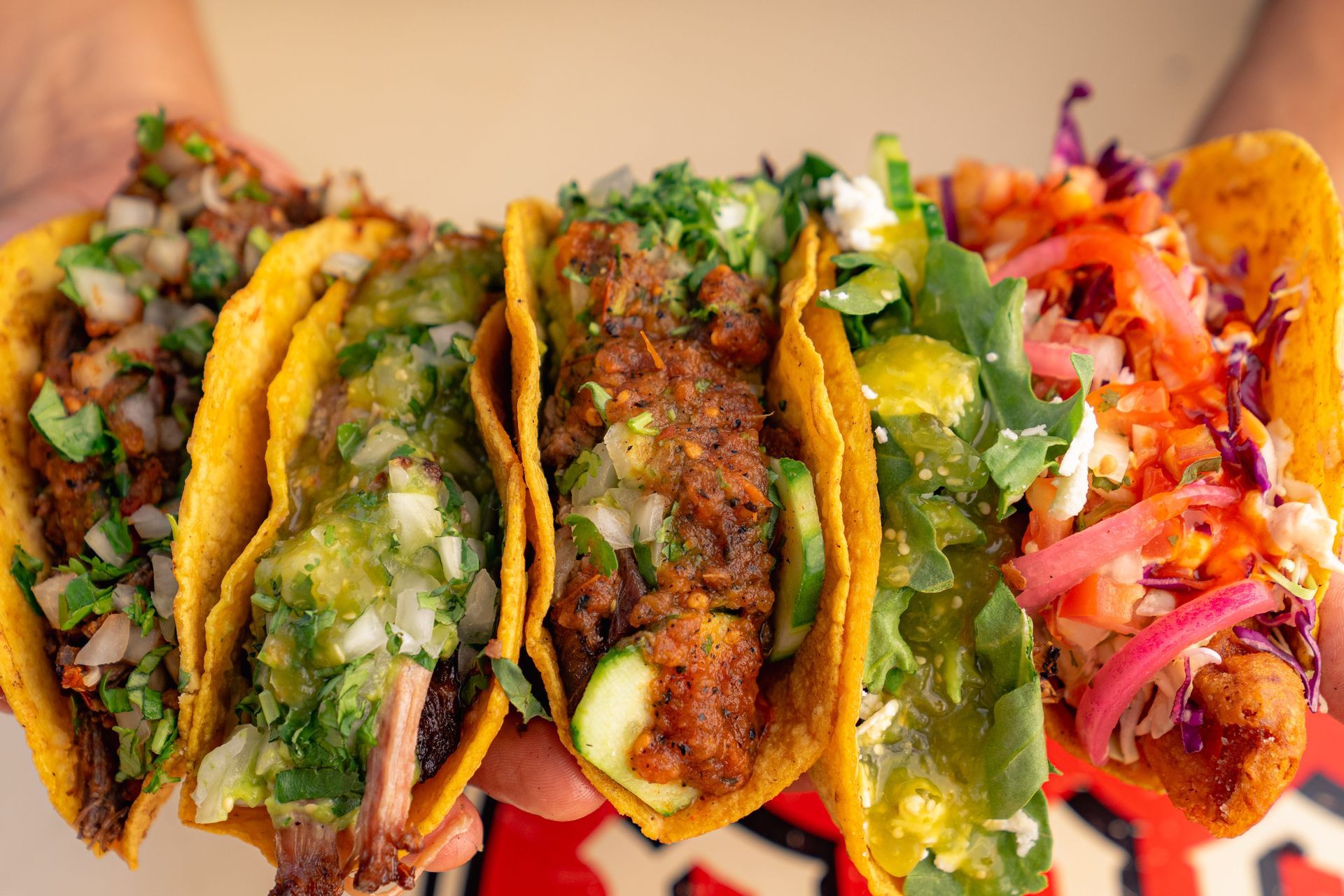 Five colorful tacos held up, each with different fillings and toppings, close-up shot.