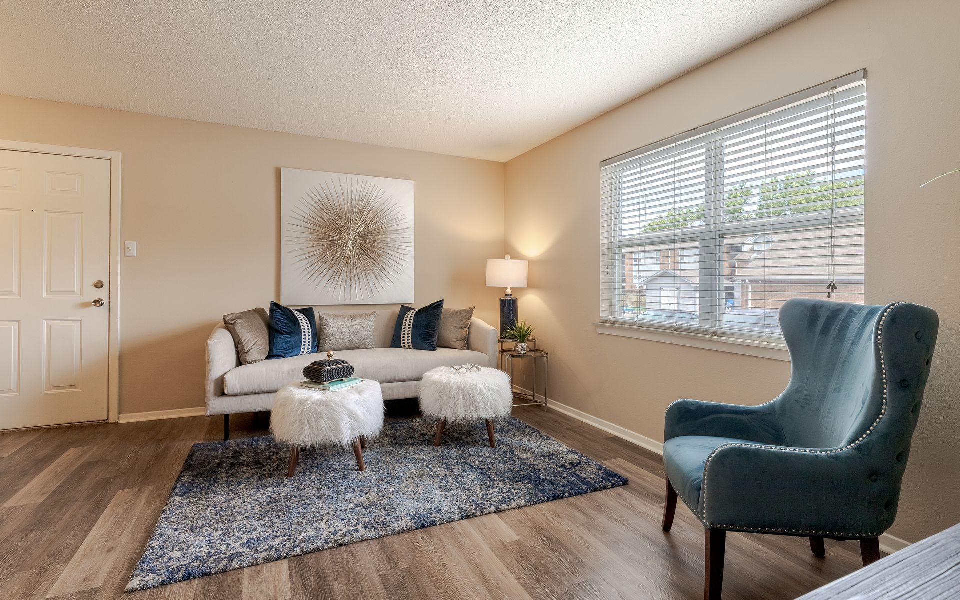 Living room with couch, teal chair, and window with blinds; beige walls, wood floors.
