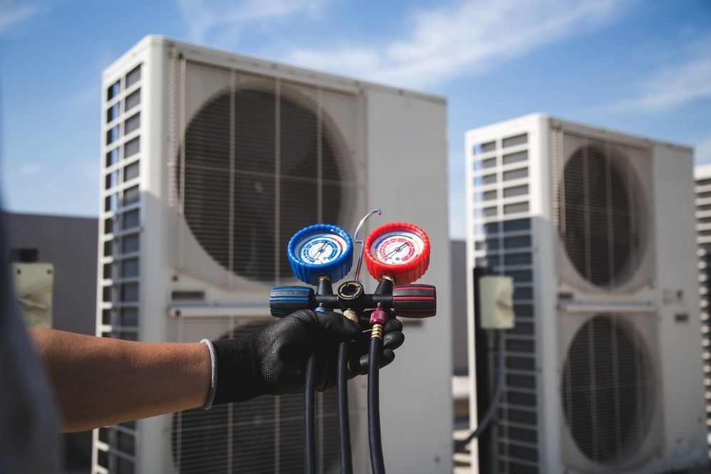 A technician in a black glove holds a blue and red HVAC manifold gauge set in front of outdoor air conditioning units.