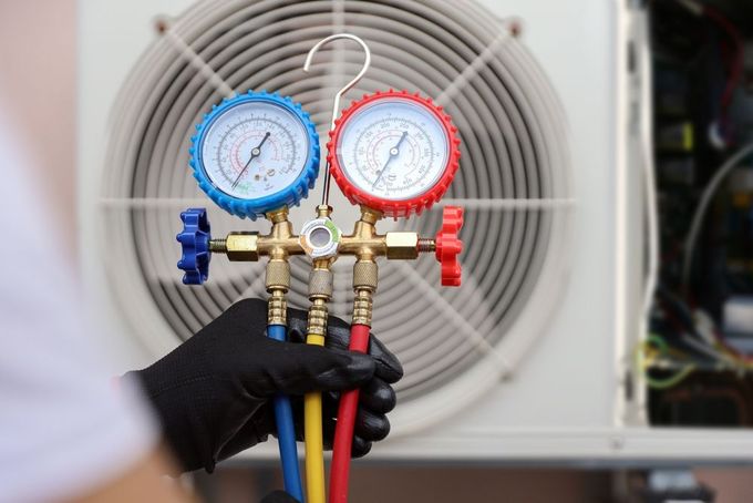 A technician in black gloves holds an HVAC manifold gauge set in front of an outdoor air conditioning unit.