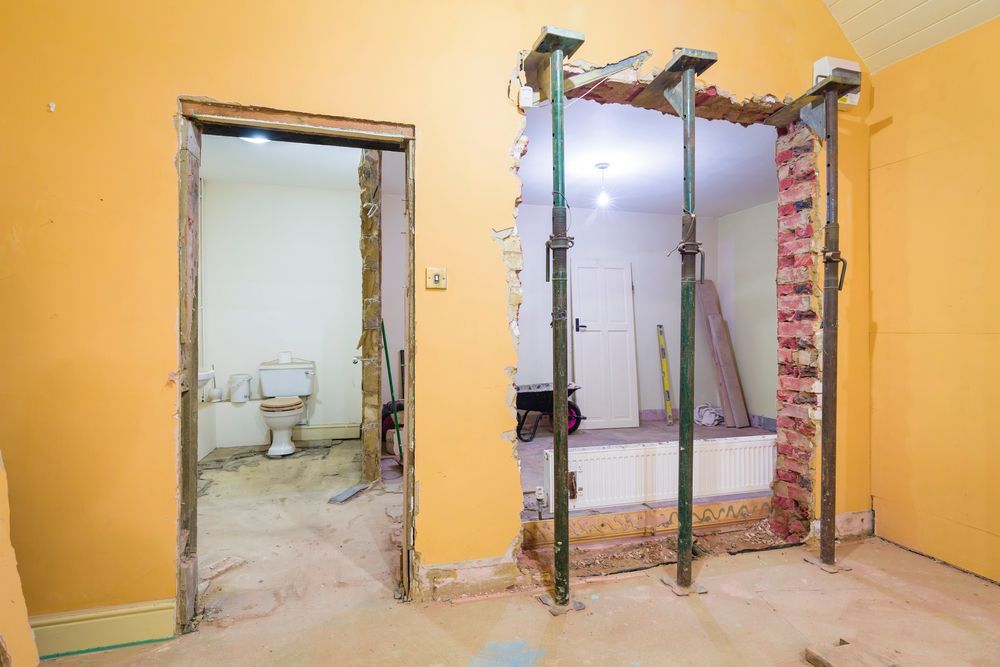 Interior construction site showing a wall opening supported by three steel adjustable props, beside a door to a bathroom.