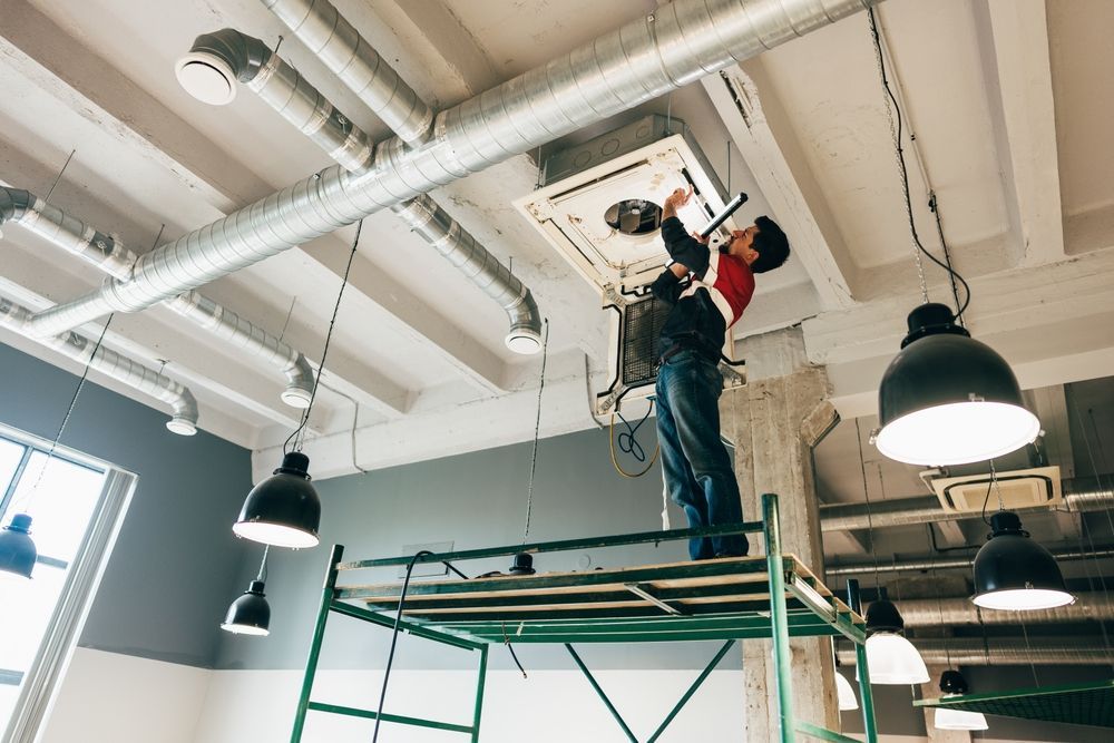A worker stands on scaffolding to repair a ceiling-mounted HVAC unit in an industrial-style room with exposed ductwork.