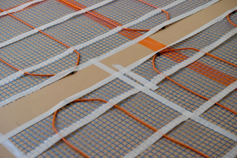 An orange electric radiant heating cable woven through a white mesh mat on a gray concrete subfloor.