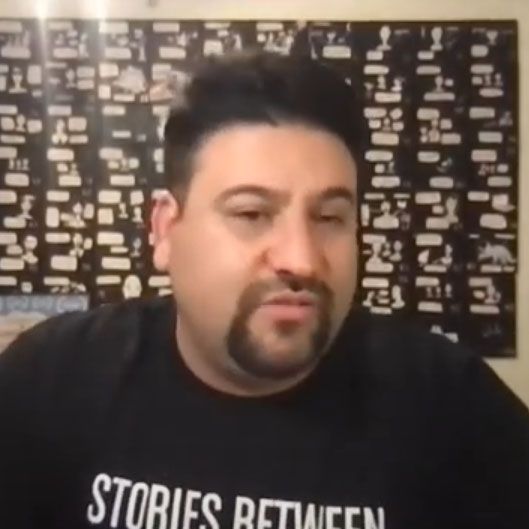 A man with a beard is wearing a black shirt that says stories between