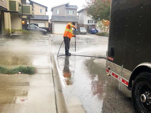Man in orange vest pressure washing a wet residential street next to a trailer.