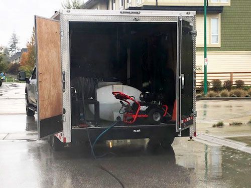 A trailer with open doors, containing a water tank and pressure washer parked on a wet street.