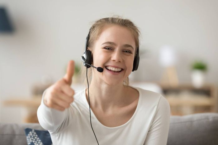Woman with headset gives a thumbs up, smiling, indoors, likely at home.