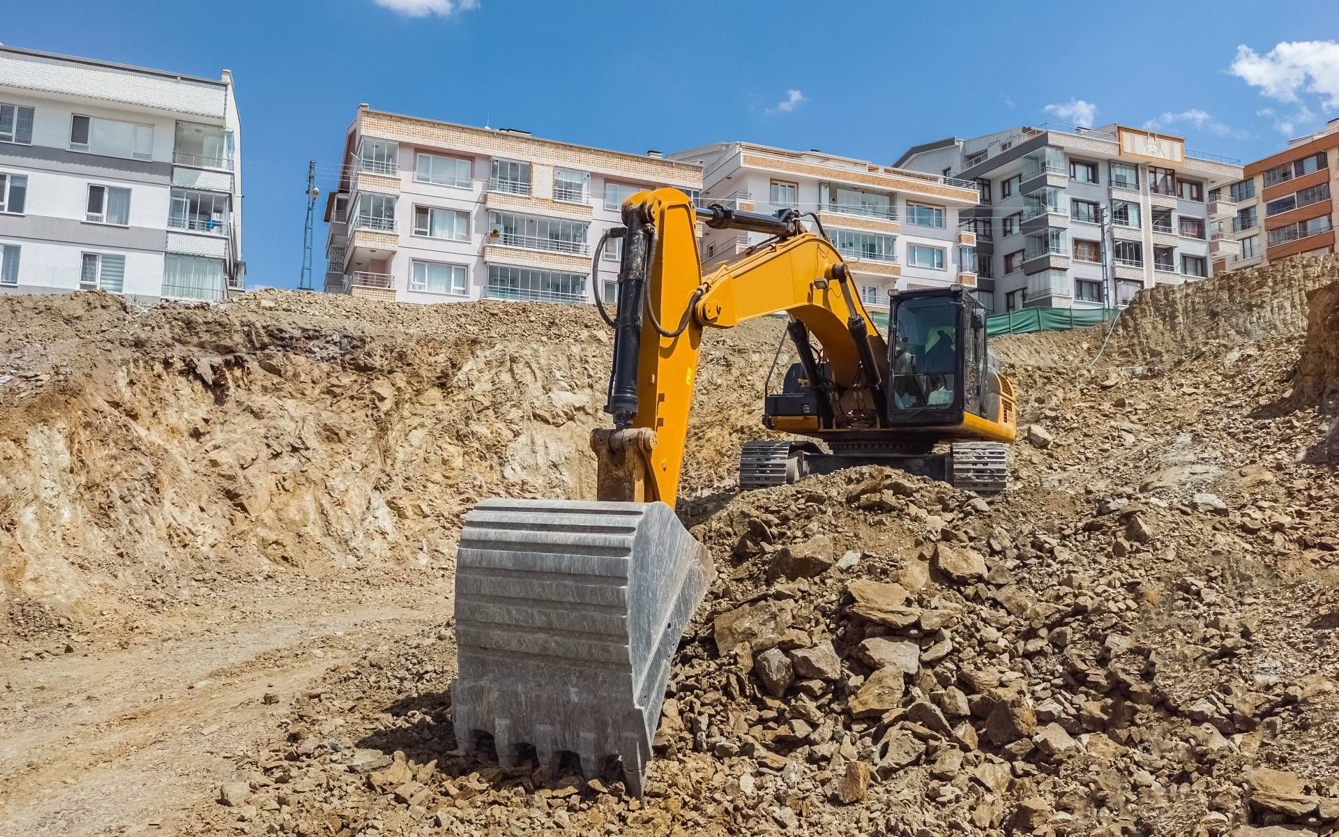 Yellow excavator on a construction site, digging into the dirt with apartment buildings in the background.
