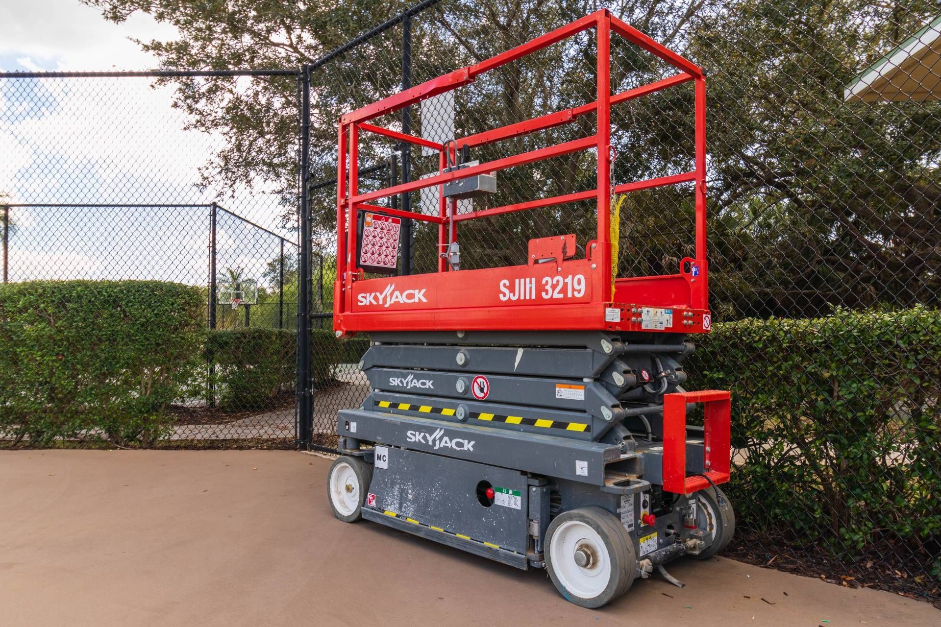 Red and grey Skyjack scissor lift on a concrete surface near a fence and hedges.