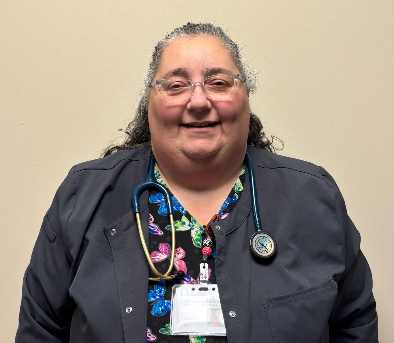 Portrait of a healthcare professional wearing dark medical scrubs and a stethoscope, standing against a plain beige background with an ID badge visible.