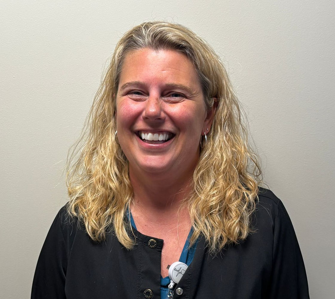 A portrait-style photograph of a person with shoulder‑length wavy blonde hair standing against a plain light-colored wall. They are wearing a dark top layered over a blue shirt, with an identification badge clipped to the front. The badge includes a photo and text, but the details are too small to read.